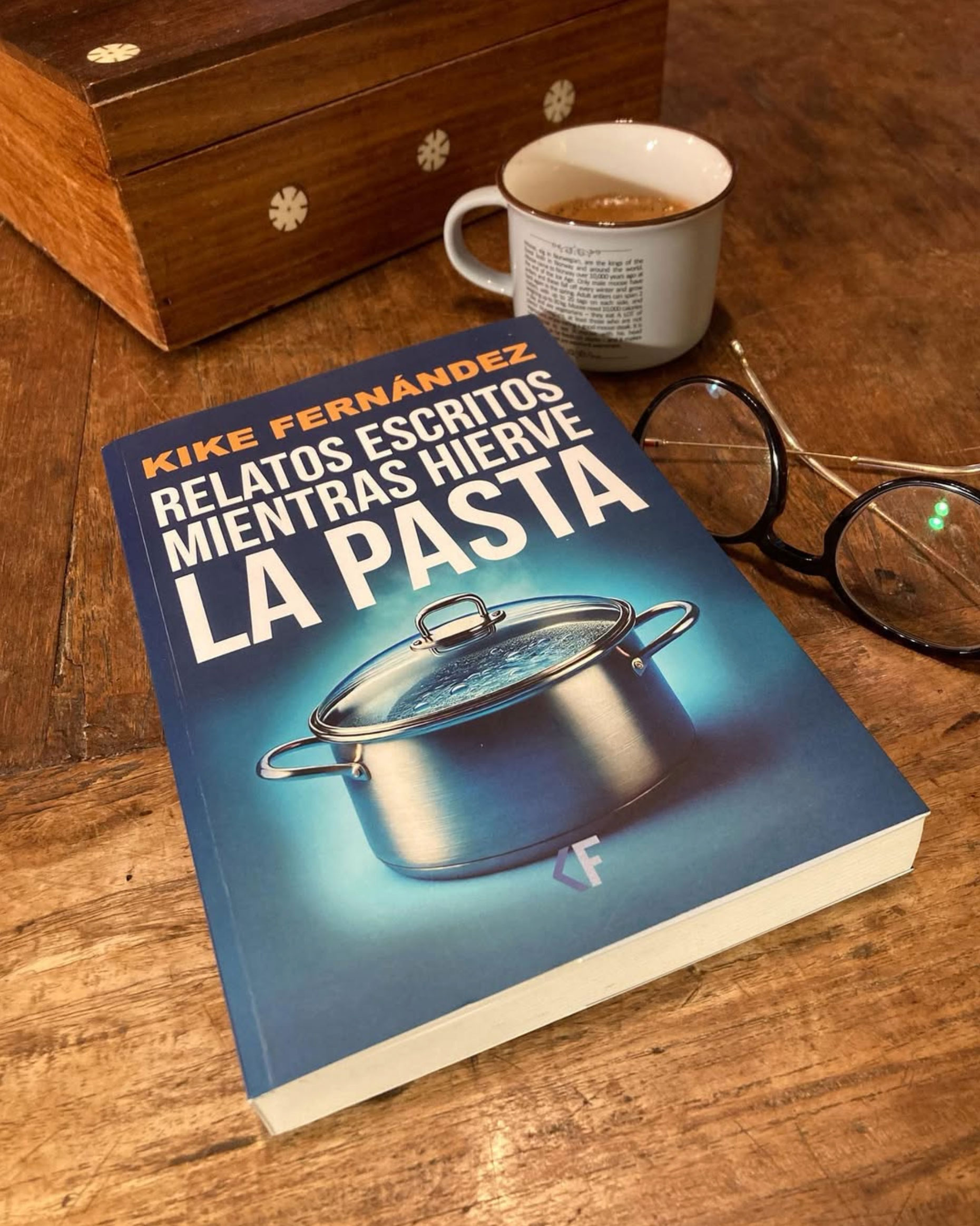 Portada del libro Relatos escritos mientras hierve la pasta, de Kike Fernández, con una olla sobre fondo azul, junto a una taza de café y unas gafas sobre una mesa de madera.