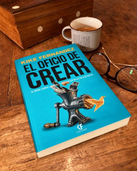 Portada del libro El oficio de crear, de Kike Fernández, con una picadora de ideas sobre fondo turquesa, junto a una taza de café y unas gafas.